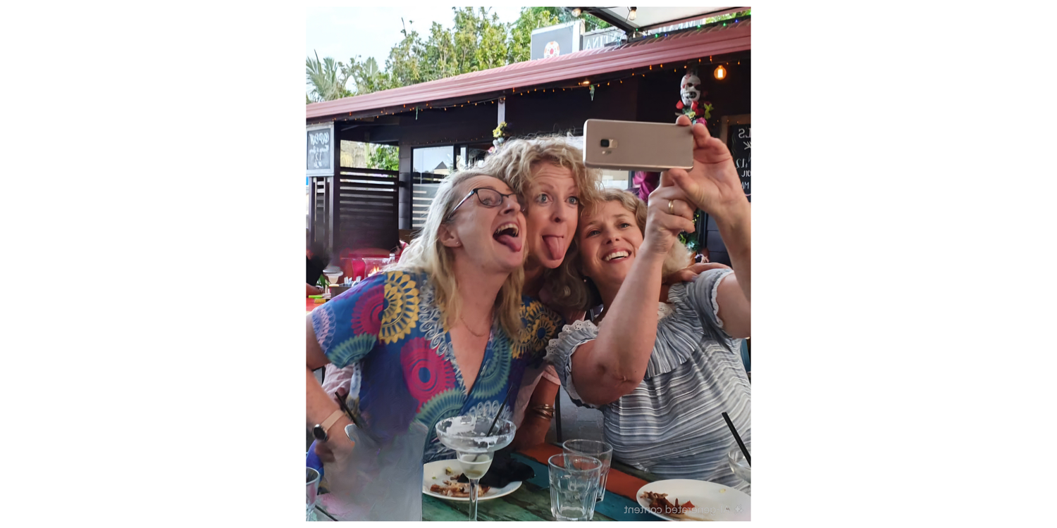 Three women taking a selfie outdoors with food and drinks on a table.
