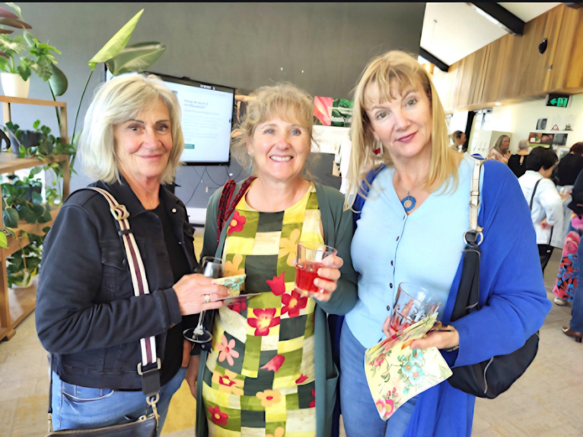 Three women standing together in a casual setting, holding drinks and smiling.